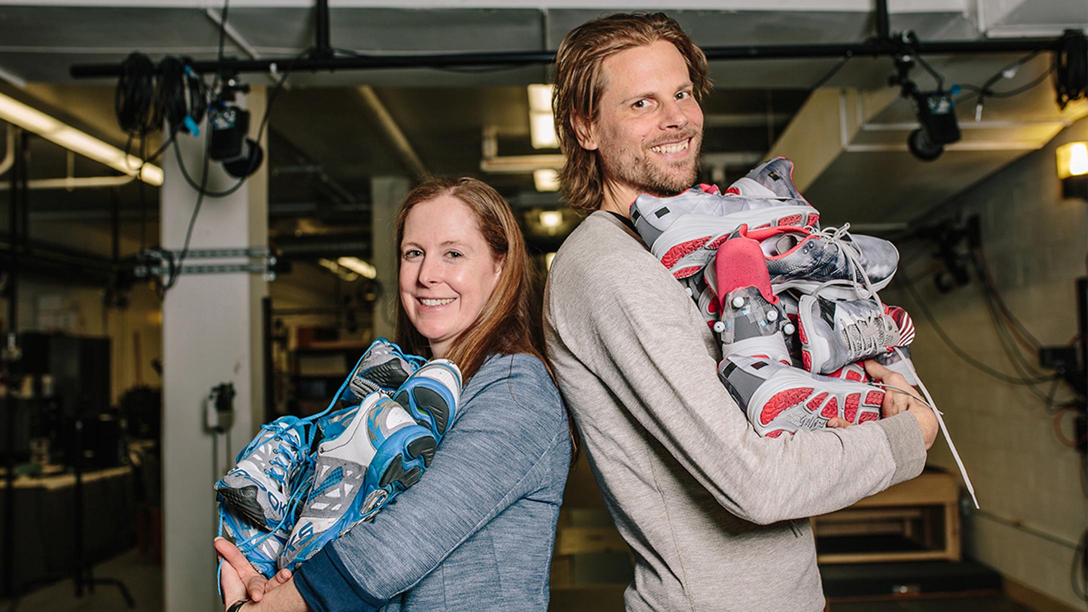 Professors Katherine Boyer and Wooter Hoogkamer of the Kinesiology lab stand back to back, each holding a large pile of athletic shoes from the testing lab.